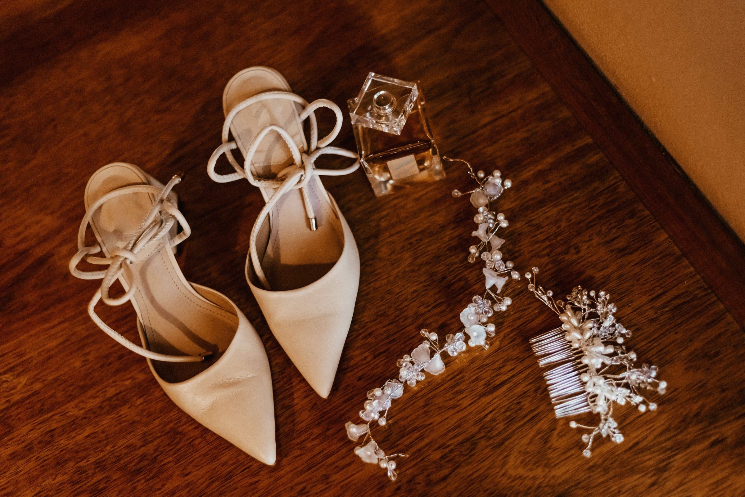 Ivory pointed bridal heels, a crystal hair vine and a small glass bottle on a wooden table